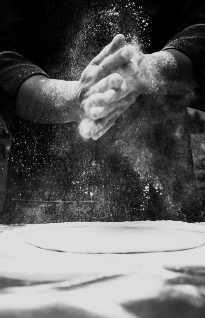 Black and white photo of a chef clapping flour in a kitchen, creating a dramatic effect.